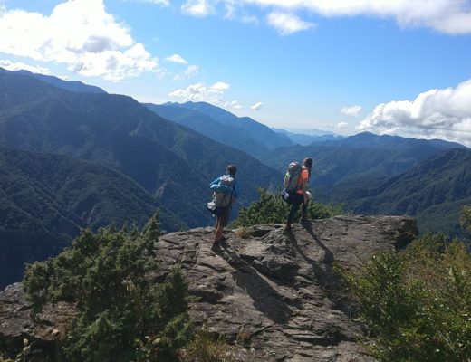 Two people on a rock, with hicking backpacks and stunning mountain scenery behind